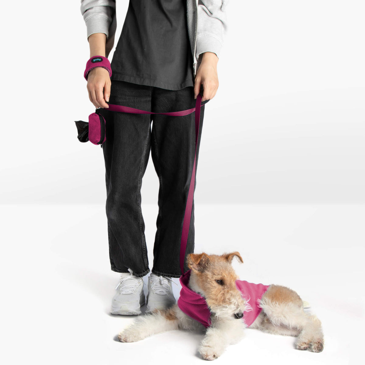 Person holding a magenta dog leash while a terrier wearing a matching magenta vest lies on the floor